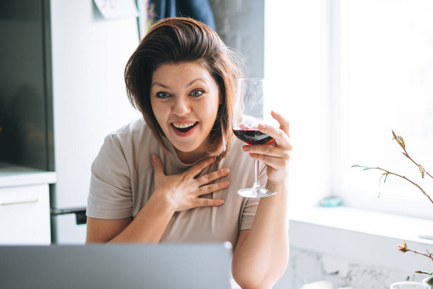 mooie gelukkig emotioneel brunette jonge vrouw plus size lichaam positief met glas wijn met video oproep op laptop in het huis - Foto, afbeelding
