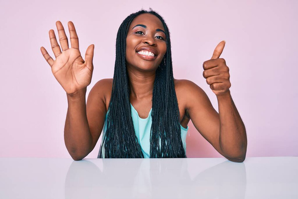 Saçları günlük kıyafetlerle örülü Afrikalı Amerikalı bir kadın masanın üzerinde oturuyor ve 6 numaralı parmaklarını göstererek gülümsüyor ve mutlu..  - Fotoğraf, Görsel
