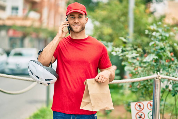Young caucasian deliveryman talking on the smartphone and holding delivery paper bag at the city. - Photo, Image