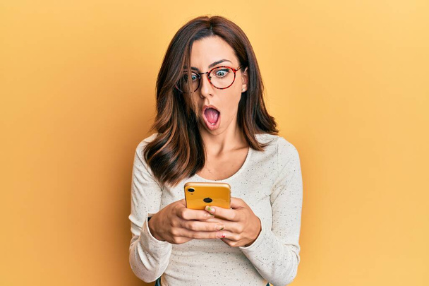 Jeune femme brune utilisant un smartphone sur fond jaune effrayé et choqué par l'expression surprise et étonnante, la peur et le visage excité.  - Photo, image