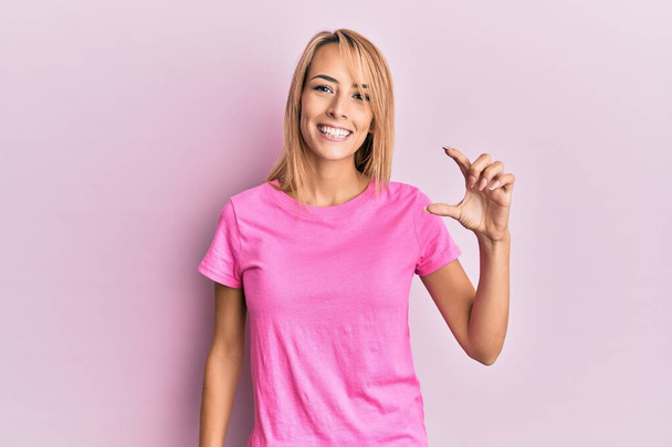 Beautiful blonde woman wearing casual pink tshirt smiling and confident gesturing with hand doing small size sign with fingers looking and the camera. measure concept.  - Fotografie, Obrázek