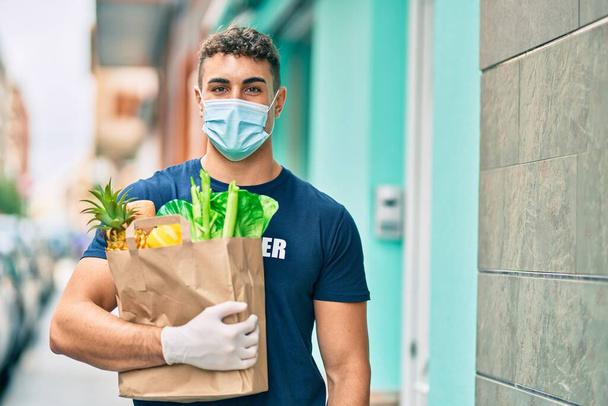 Young hispanic volunteer man wearing medical mask holding groceries at the city. - Photo, Image