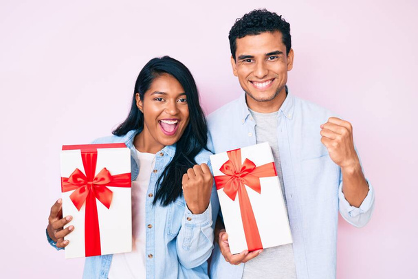 Beautiful latin young couple holding gift for anniversary celebration screaming proud, celebrating victory and success very excited with raised arms  - Foto, Bild