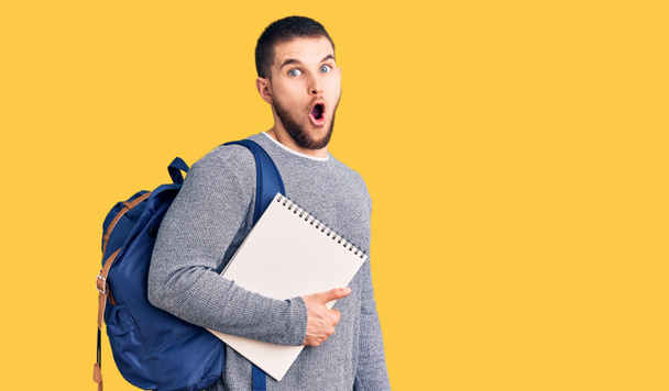 Jonge knappe man draagt student rugzak met notebook bang en verbaasd met open mond voor verrassing, ongeloof gezicht  - Foto, afbeelding