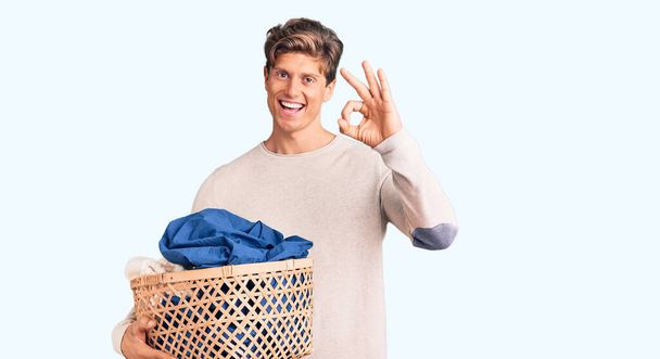 Young handsome man holding laundry basket doing ok sign with fingers, smiling friendly gesturing excellent symbol  - Foto, Imagen