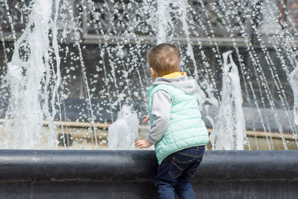 Odesa, Ukraine - 2020/01/05: Little boy near the fountain. Water jets, splashes and drops from the fountain on a hot summer day. - Photo, Image