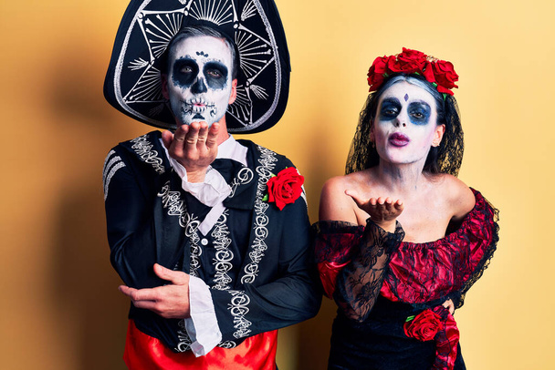 Young couple wearing mexican day of the dead costume over yellow looking at the camera blowing a kiss with hand on air being lovely and sexy. love expression.  - Fotoğraf, Görsel