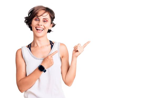 Красивая молодая женщина с короткими волосами в повседневной спортивной одежде улыбается и смотрит в камеру, указывая двумя руками и пальцами в сторону.  - Фото, изображение