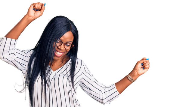 若いアフリカ系アメリカ人女性がカジュアルな服と眼鏡を身に着けて幸せと陽気に踊り、笑顔でカジュアルで自信を持って音楽を聴く  - 写真・画像