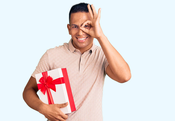 Joven hombre latino guapo sosteniendo regalo sonriendo feliz haciendo ok signo con la mano en el ojo mirando a través de los dedos  - Foto, imagen