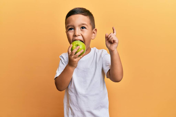 Suloinen latino taapero hymyilee onnellinen syöminen vihreä omena etsii kameraan yli eristetty keltainen tausta. - Valokuva, kuva