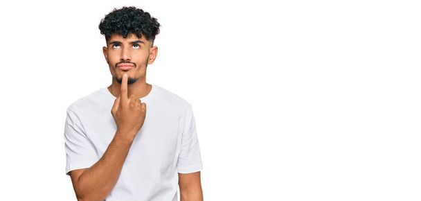 Hombre árabe joven con camiseta blanca casual pensando concentrado en la duda con el dedo en la barbilla y mirando hacia arriba preguntándose  - Foto, Imagen