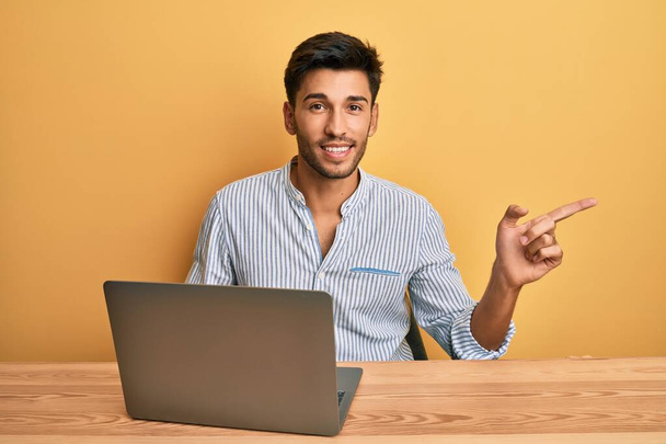 Молодой красивый мужчина работает в офисе с ноутбуком улыбается счастливо указывая рукой и пальцем в сторону  - Фото, изображение