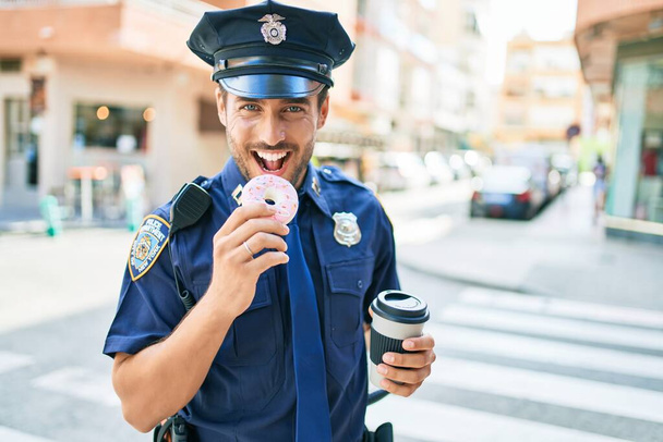 Joven policía hispano guapo vistiendo uniforme de policía sonriendo feliz. Comer rosquilla y beber una taza de café para llevar en la calle de la ciudad. - Foto, imagen