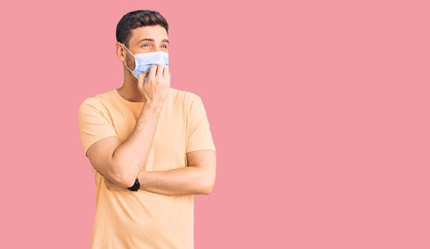 Schöner junger Mann mit Bär, der eine medizinische Maske gegen Coronavirus trägt, sieht gestresst und nervös aus, die Hände auf den Mund beißenden Nägeln. Angstproblem.  - Foto, Bild