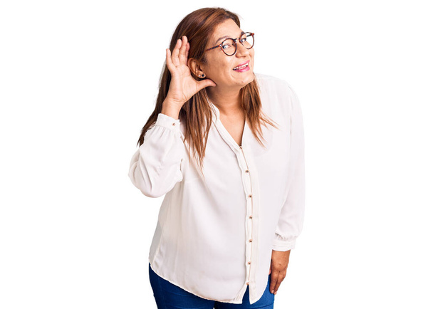 Középkorú latin nő alkalmi ruhákat és szemüveget visel, kéztől fülig mosolyog, és egy pletykát vagy pletykát hallgat. süketség fogalma.  - Fotó, kép
