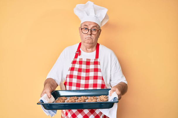 Idősebb ősz hajú férfi, pékegyenruhában, házi készítésű sütikkel a kezében, tudatlan és zavaros arckifejezéssel. kétség.  - Fotó, kép