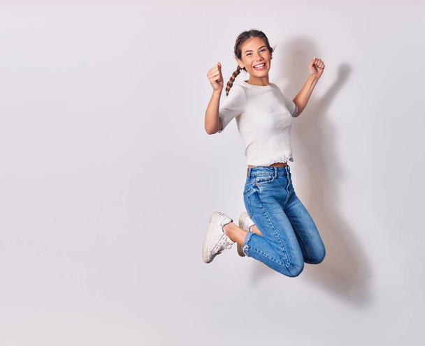 Young beautiful woman with braids wearing casual clothes smiling happy. Jumping with smile on face doing winner sign with fists up over isolated white background - Fotó, kép