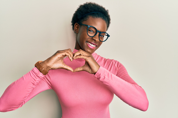 Jong Afrikaans Amerikaans meisje draagt casual kleding en glazen glimlachen in liefde doen hart symbool vorm met handen. romantisch concept.  - Foto, afbeelding