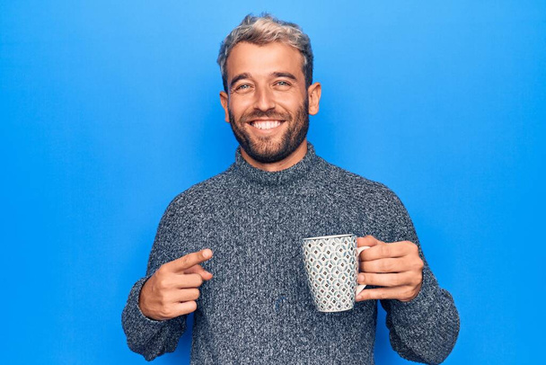 Sakallı, genç, sarışın bir adam izole edilmiş mavi arka planda bir kupa kahve içerken mutlu bir şekilde eli ve parmağı ile işaret ediyor. - Fotoğraf, Görsel