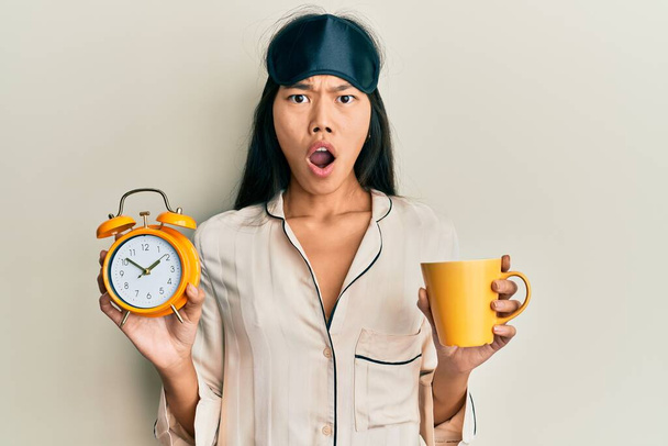 Young chinese woman wearing sleep mask and robe holding alarm clock and cup of coffee afraid and shocked with surprise and amazed expression, fear and excited face.  - Photo, Image