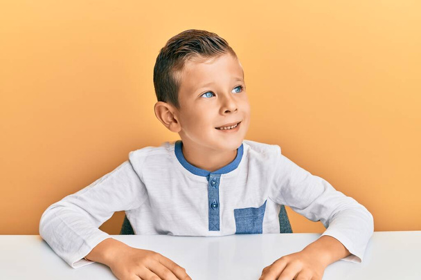 Entzückendes kaukasisches Kind in lässiger Kleidung, das auf dem Tisch sitzt und zur Seite schaut, mit einem Lächeln im Gesicht, natürlichem Ausdruck. Lachen selbstbewusst.  - Foto, Bild