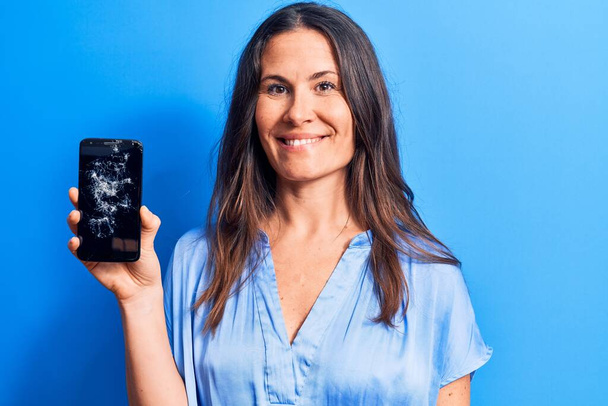 Giovane bella donna bruna in possesso di smartphone rotto mostrando schermo rotto guardando positivo e felice in piedi e sorridendo con un sorriso fiducioso mostrando i denti - Foto, immagini
