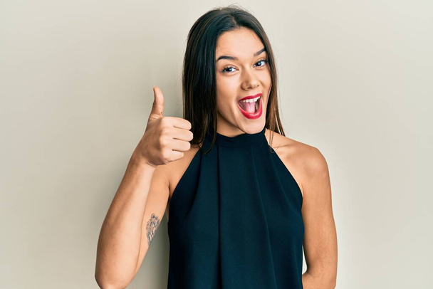 Молодая латиноамериканка носит повседневный стиль с рубашкой без рукавов улыбается счастливой и позитивной, большой палец вверх делает отлично и знак одобрения  - Фото, изображение