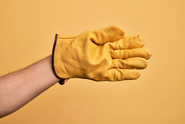 Hand eines kaukasischen jungen Mannes mit Gärtnerhandschuh über isoliertem gelben Hintergrund, der sich streckt und mit offener Hand nach Handschlag greift, zeigt Handfläche - Foto, Bild