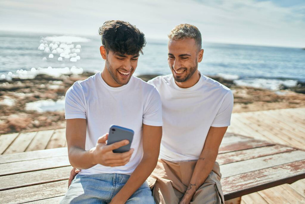 Молода гей-пара посміхається щасливий, використовуючи смартфон на пляжі
. - Фото, зображення