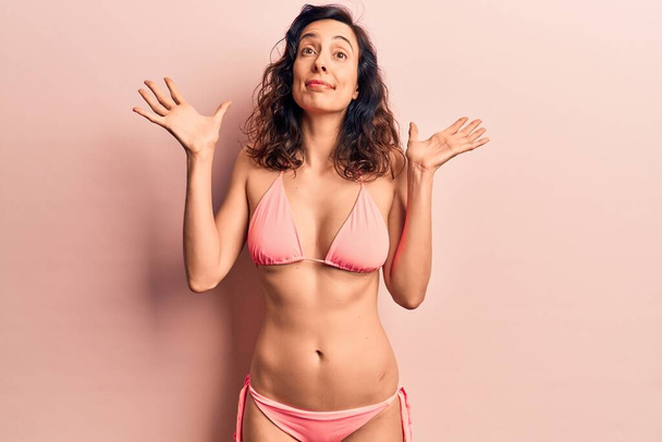Young beautiful hispanic woman wearing bikini clueless and confused with open arms, no idea and doubtful face.  - Foto, afbeelding