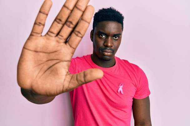 bel homme noir portant un ruban de cancer rose attaché sur la chemise avec la main ouverte faisant stop signe avec expression sérieuse et confiante, geste de défense  - Photo, image