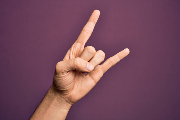 Hand eines kaukasischen jungen Mannes mit Fingern vor isoliertem lila Hintergrund, gestikulierend als Rock "n" Roll-Symbol, mit obszöner Hörner-Geste - Foto, Bild
