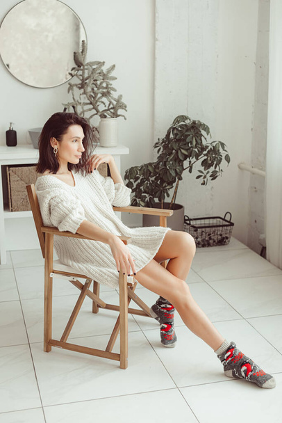 A young beautiful girl, a brunette, in a bright room during the day, interiors from light ECO furniture, the girl is wearing a white sweater and socks - Foto, immagini