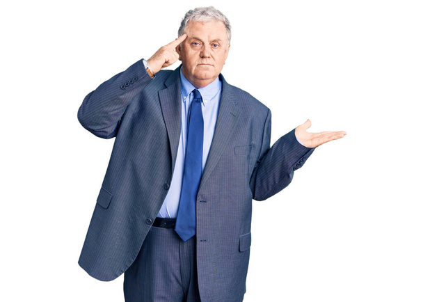 Älterer grauhaariger Mann in Business-Jacke verwirrt und genervt von offener Handfläche, die Kopierraum zeigt und mit dem Finger auf die Stirn zeigt. Denken Sie darüber nach.  - Foto, Bild