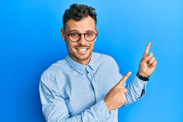 Nuori latinomies yllään rento vaatteita ja lasit hymyillen ja katsoen kameraa osoittaa kahdella kädellä ja sormella sivulle.  - Valokuva, kuva