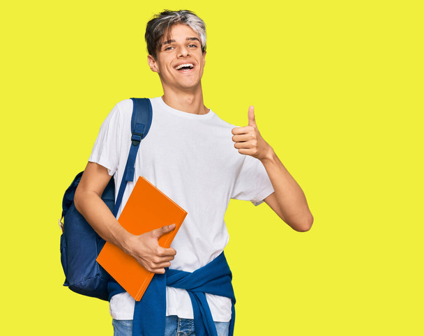 Joven hispano vistiendo mochila de estudiante y sosteniendo libros sonriendo feliz y positivo, pulgar hacia arriba haciendo excelente y signo de aprobación  - Foto, imagen
