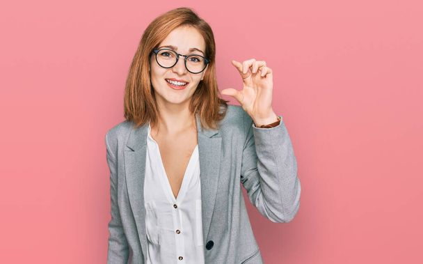 ビジネススタイルとメガネを身に着けている若い白人女性は、指を見てカメラで小さなサイズの看板を行う手で笑顔と自信を持ってジェスチャー。概念を測定し.  - 写真・画像