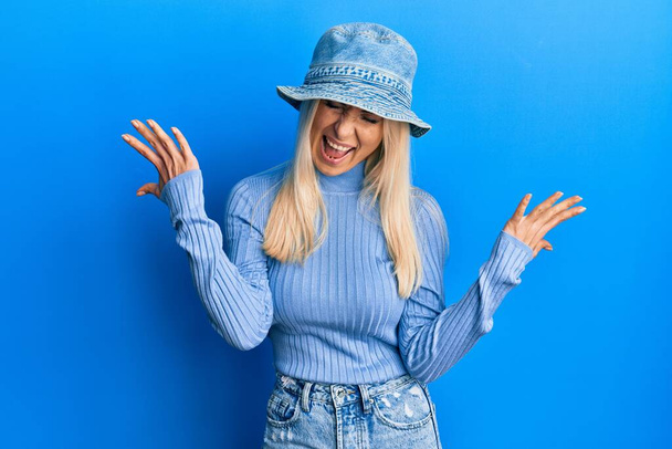 Mladá blondýnka v neformálním denimovém klobouku oslavuje šílenost a šílenství z úspěchu se zdviženýma a zavřenýma očima a vzrušeně křičí. koncept vítěze  - Fotografie, Obrázek