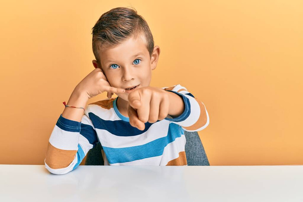 Adorable caucasian kid wearing casual clothes sitting on the table smiling doing talking on the telephone gesture and pointing to you. call me.  - Photo, Image