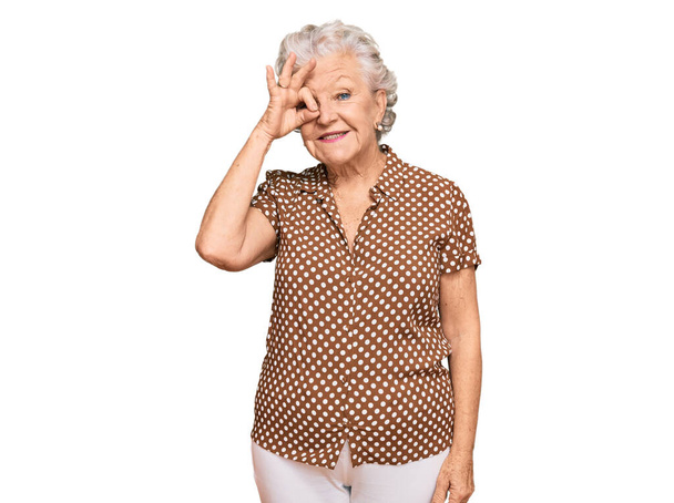 Senior mulher de cabelos grisalhos vestindo roupas casuais sorrindo feliz fazendo ok sinal com a mão no olho olhando através dos dedos  - Foto, Imagem