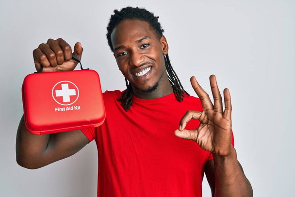 Africký Američan s copánky drží lékárničku a prsty drží ceduli, přátelsky se usmívá a gestikuluje znamenitým symbolem  - Fotografie, Obrázek