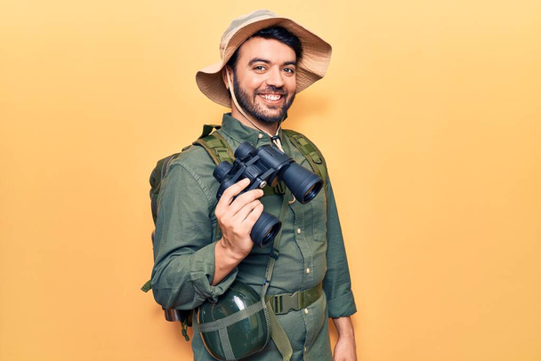 Jeune homme hispanique portant des vêtements d'explorateur tenant des jumelles regardant positif et heureux debout et souriant avec un sourire confiant montrant des dents  - Photo, image