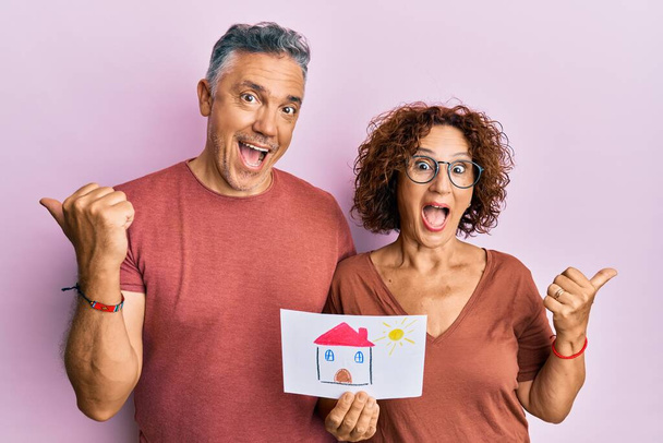 красивая пара среднего возраста держит дом рисовать покупая дома для будущего указывая большой палец вверх в сторону улыбаясь счастливым с открытым ртом  - Фото, изображение