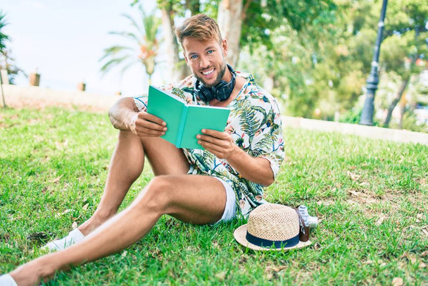 Schöner kaukasischer Mann mit Sommermütze und Blumenhemd, der glücklich auf dem Gras sitzt und ein Buch liest - Foto, Bild