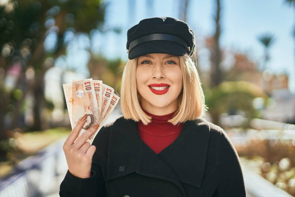 Молодая блондинка улыбается счастливо держа Соединенное Королевство фунтов банкноты в городе. - Фото, изображение