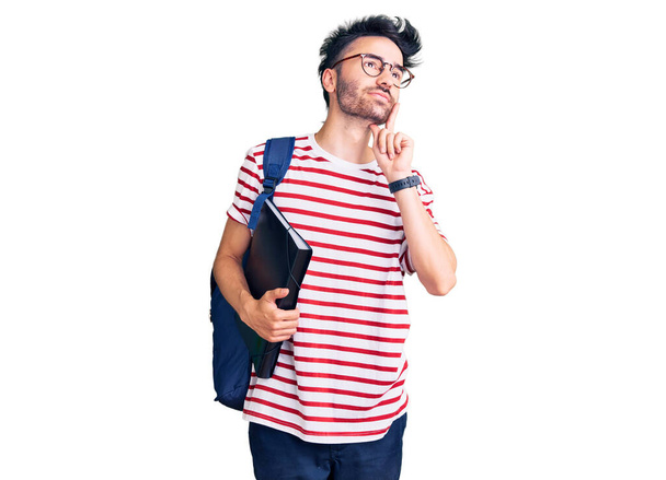 Jeune homme hispanique portant un sac à dos d'étudiant et des lunettes tenant un cartable visage sérieux réfléchissant à la question avec la main sur le menton, réfléchissant à une idée déroutante  - Photo, image