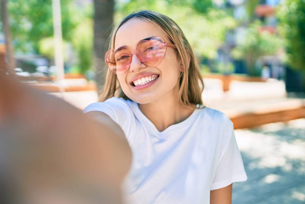 Joven hermosa rubia caucásica sonriendo feliz al aire libre en un día soleado usando gafas de sol en forma de corazón tomando una foto selfie - Foto, Imagen
