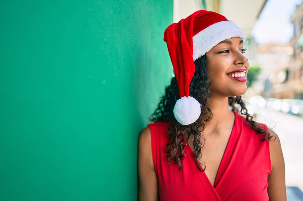 Νεαρή Αφροαμερικανή που φοράει χριστουγεννιάτικο καπέλο χαμογελώντας χαρούμενη ακουμπισμένη στον τοίχο. - Φωτογραφία, εικόνα