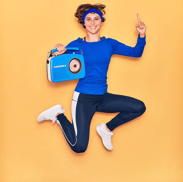 Young beautiful sportswoman listening to music smiling happy. Jumping with smile on face holding vintage radio over isolated yellow background. - Foto, imagen